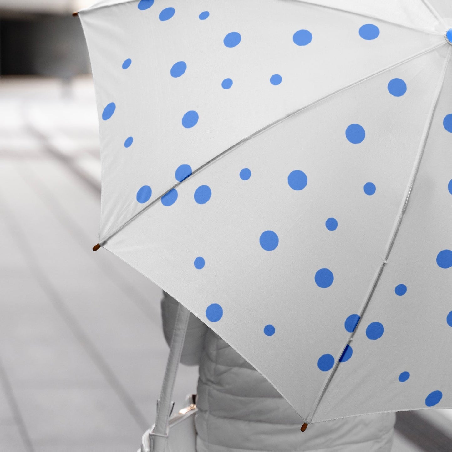 A person holds an open white umbrella with blue polka dots, walking on a grey sidewalk.