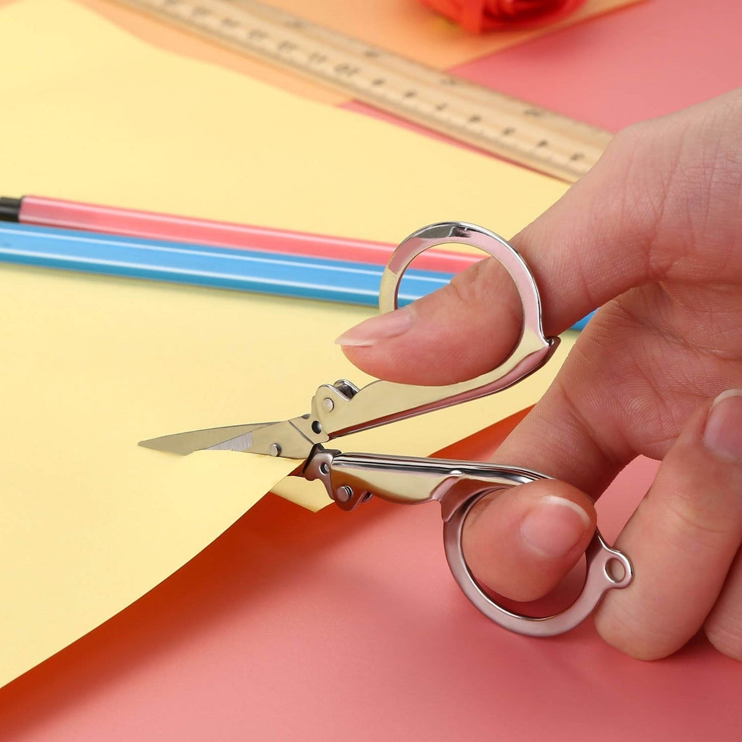 A hand uses open silver folding scissors to cut yellow paper, with a ruler and pencils in the background.