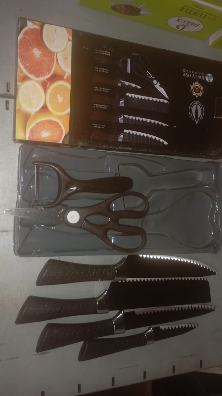 Six-piece black serrated knife set with peeler and scissors in their packaging, next to a box with fruit images.