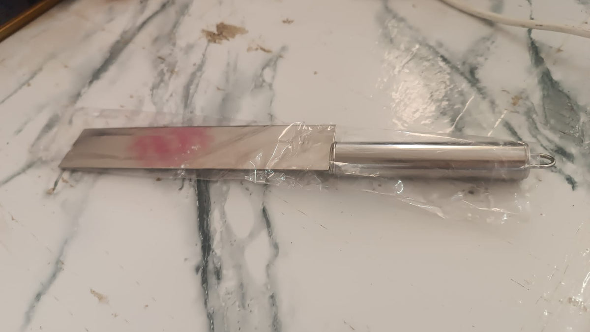 A stainless steel cleaver knife, partially wrapped in clear plastic, rests on a white marble surface with faint pink marks.