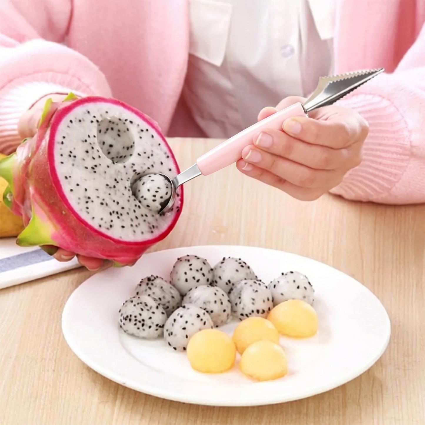 A person uses a pink-handled 2-in-1 tool to scoop dragon fruit balls, with fruit balls on a white plate.