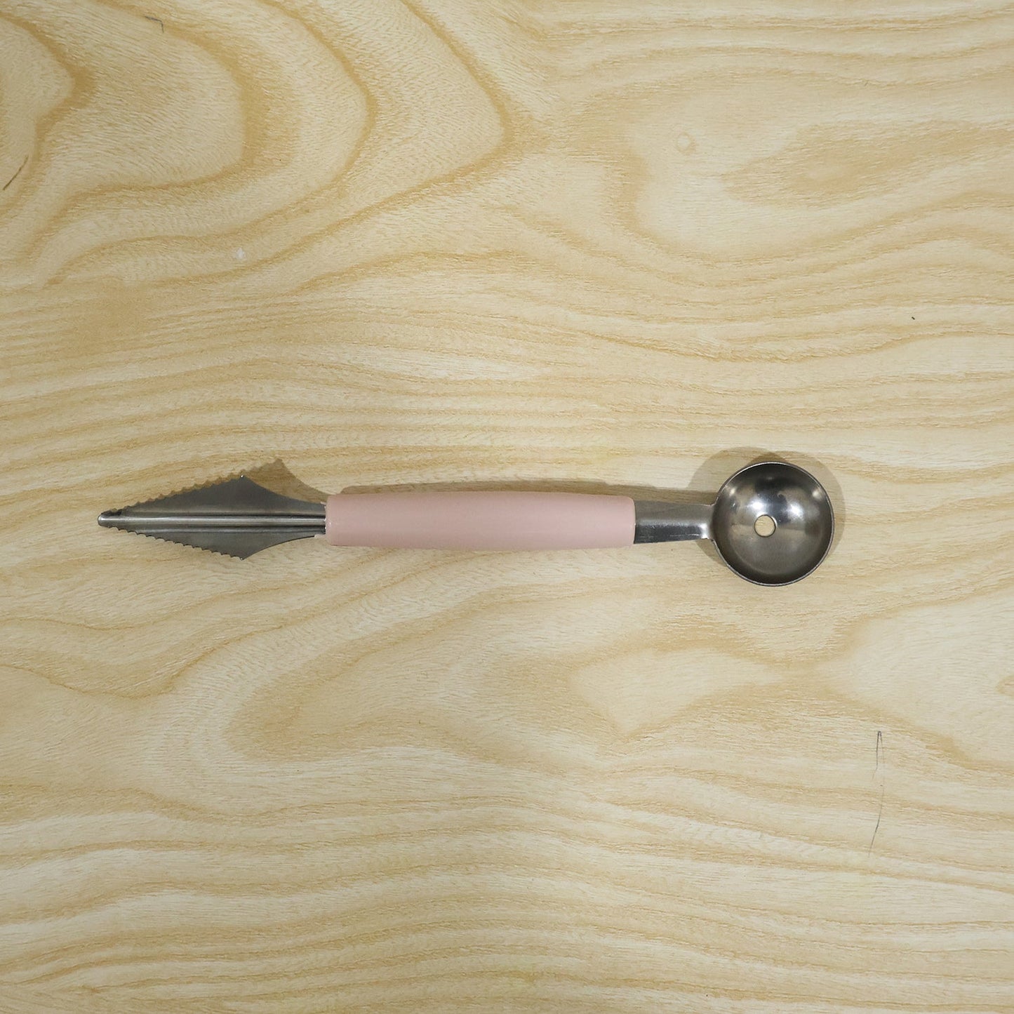 A pink-handled 2-in-1 fruit carving tool with a melon baller and a zigzag knife, lying flat on a light wood surface.