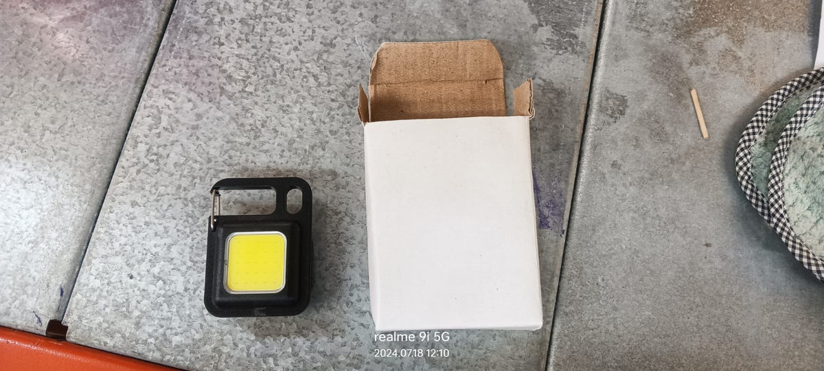 A black mini LED flashlight with a yellow light next to an open, empty white cardboard box on a textured gray surface.