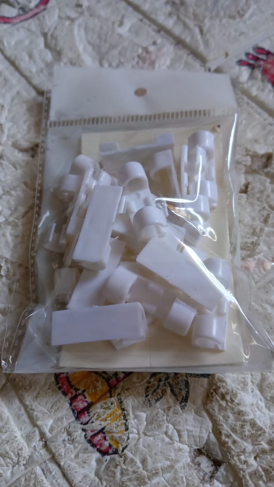 A clear plastic bag filled with numerous white plastic cable clips rests on a patterned fabric surface.