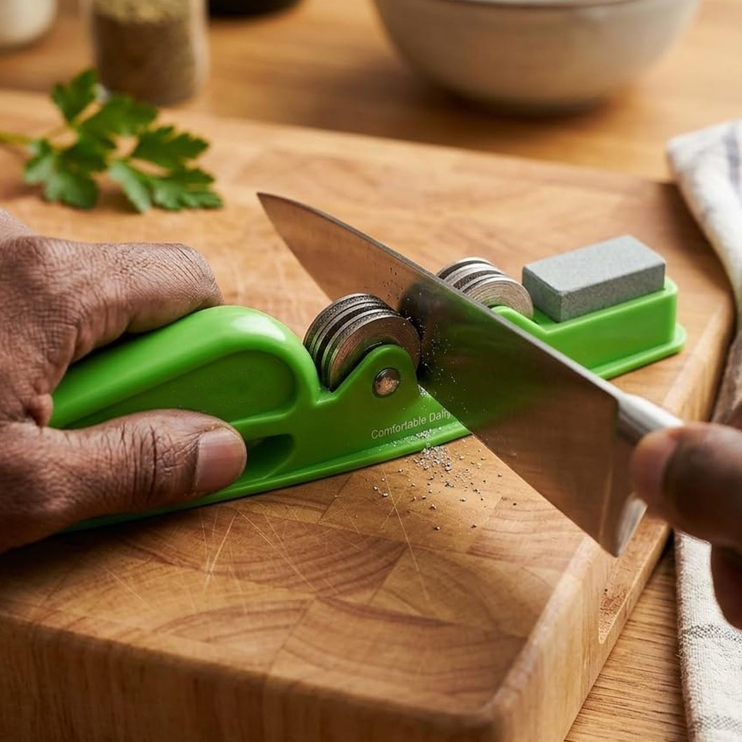A person's hands sharpening a knife with a green handheld sharpener on a wooden cutting board, with herbs and spices in the