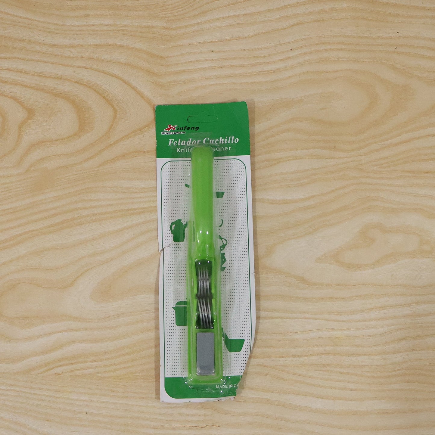 A green handheld knife sharpener in its clear plastic and green cardboard packaging, lying on a light wooden surface.