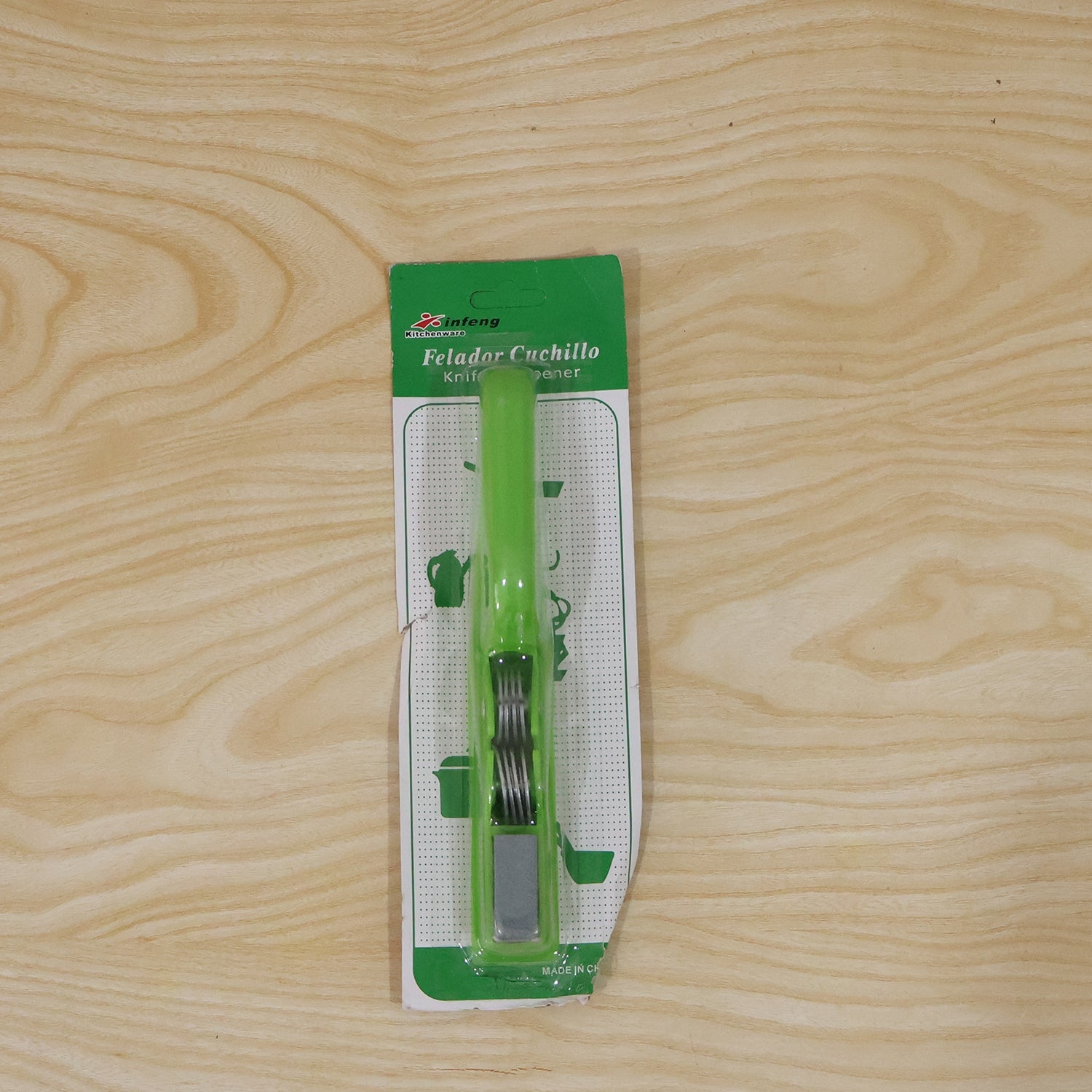 A green handheld knife sharpener in its clear plastic and green cardboard packaging, lying on a light wooden surface.