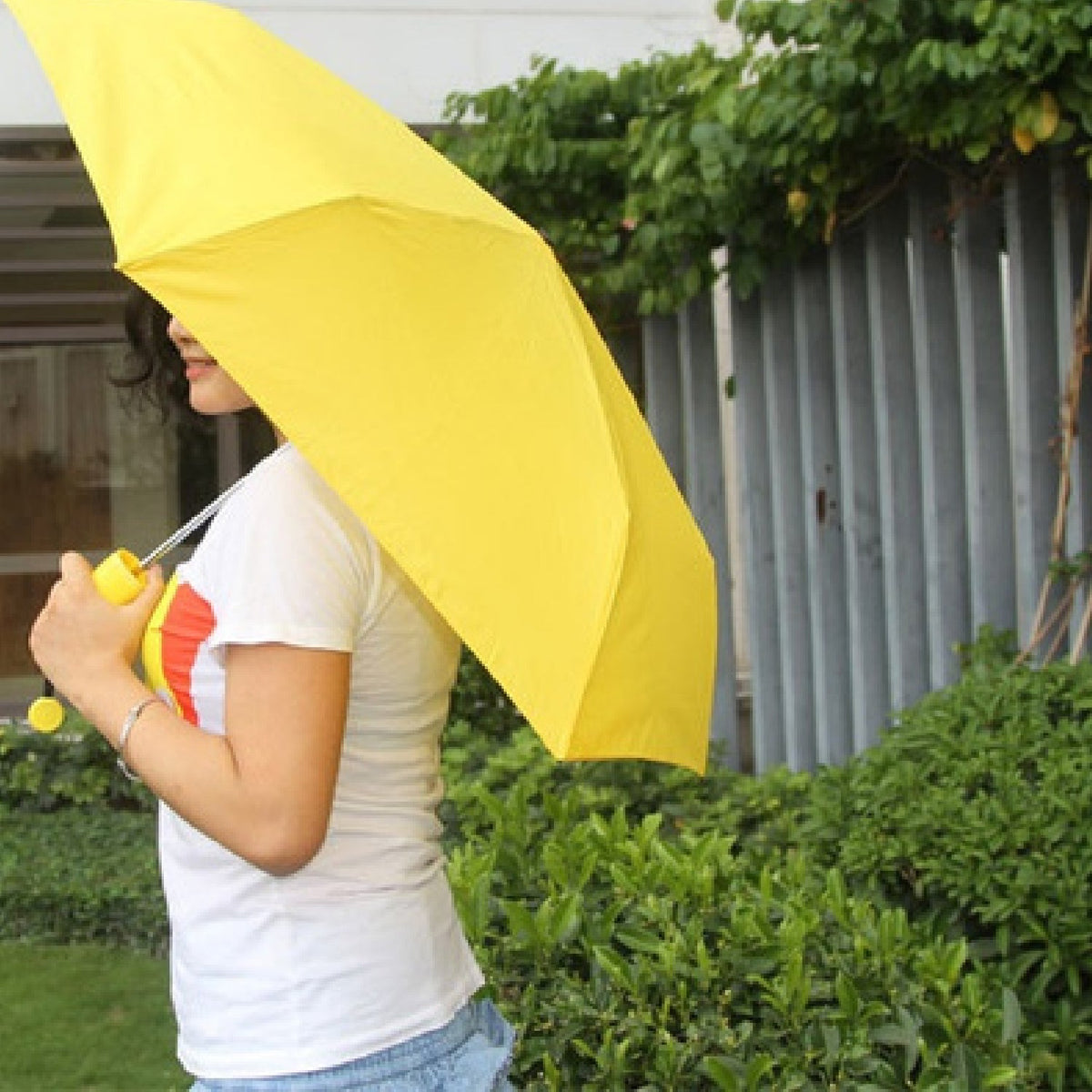 A person holds a yellow umbrella open, walking past green bushes and a fence in a garden setting.