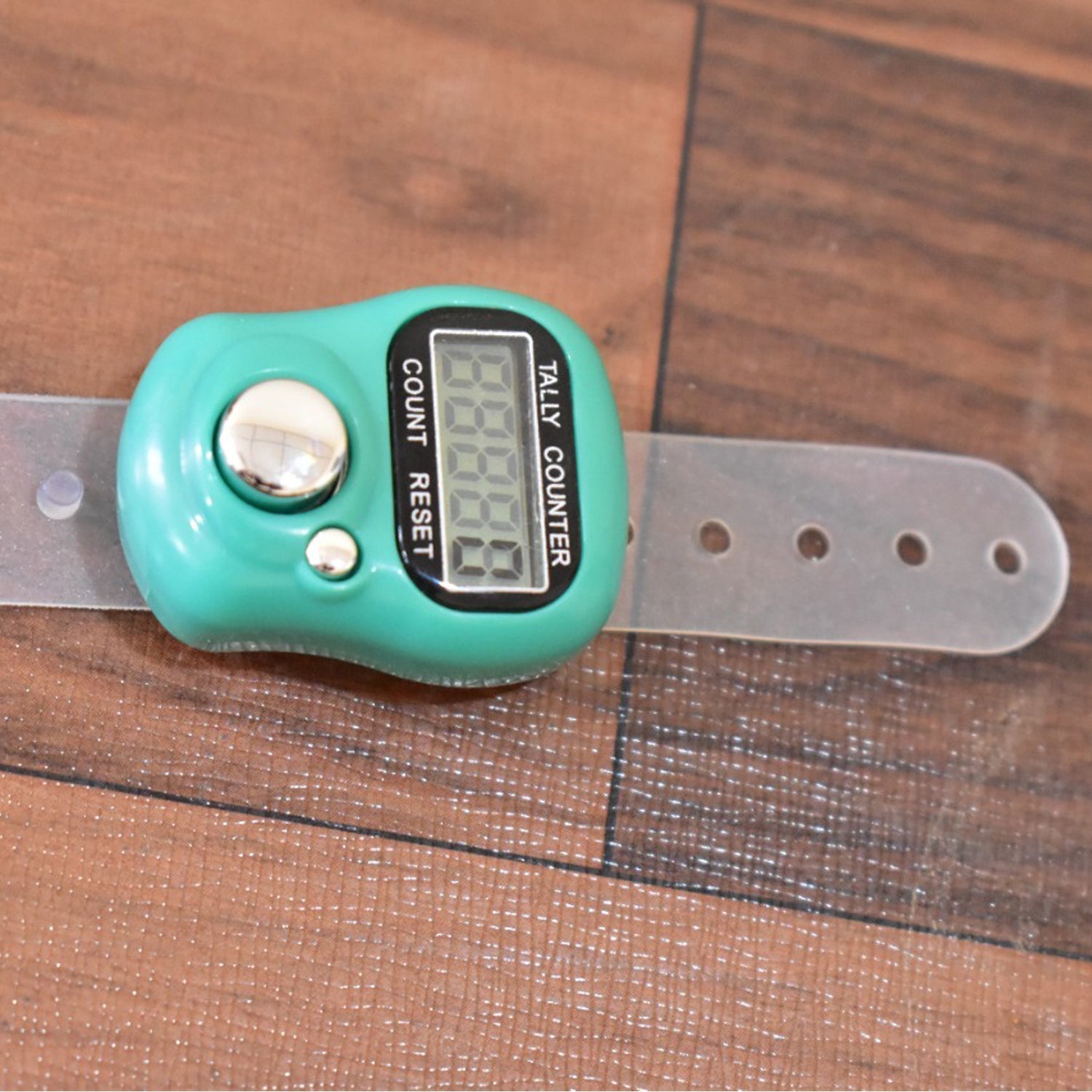A green digital tally counter with a clear plastic strap lies on a brown wooden surface.