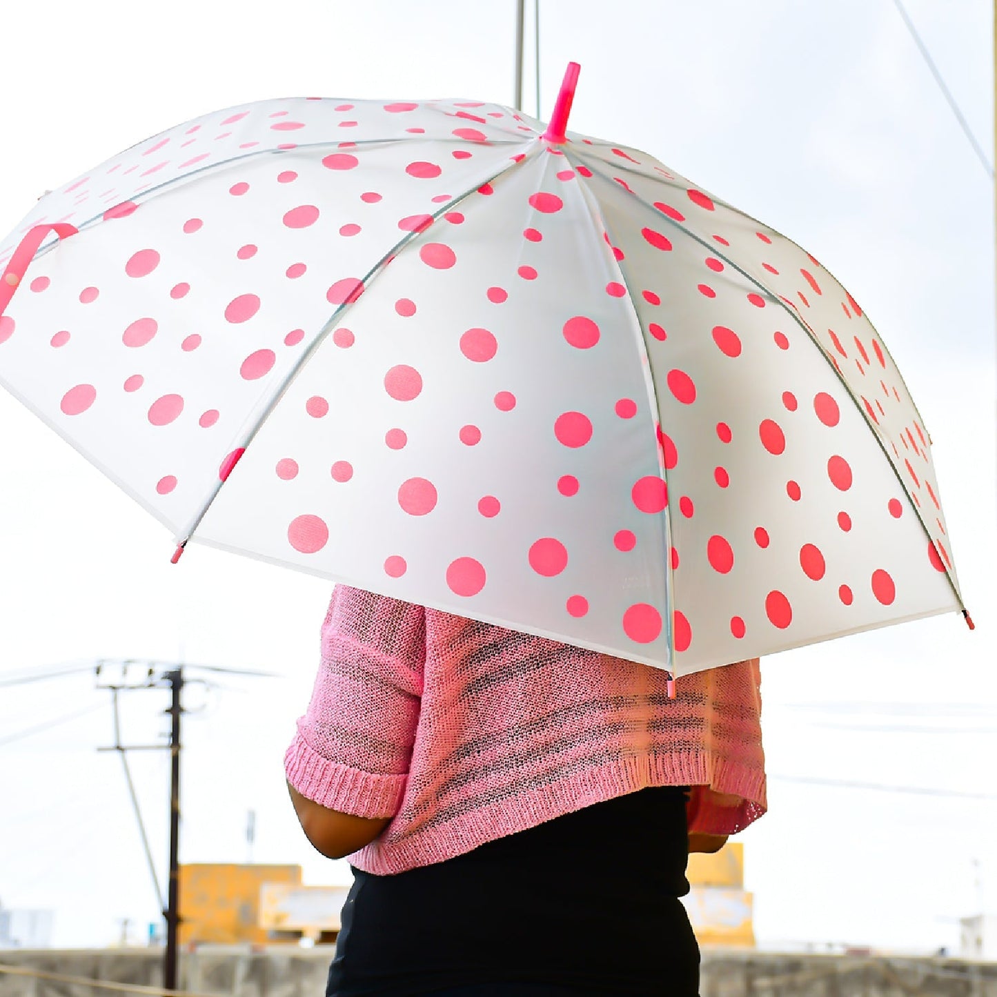 The back of a person holding an open white umbrella with pink polka dots, wearing a pink sweater.