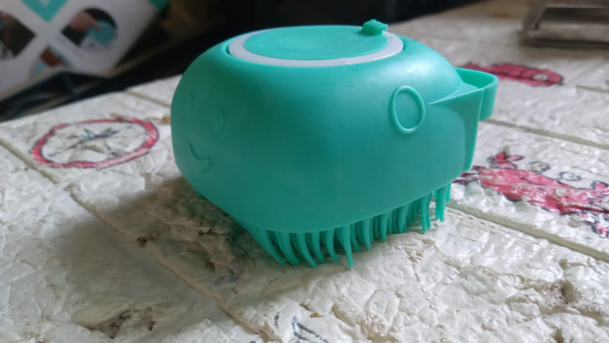 Teal silicone bath brush with shampoo dispenser on a textured white and pink surface.