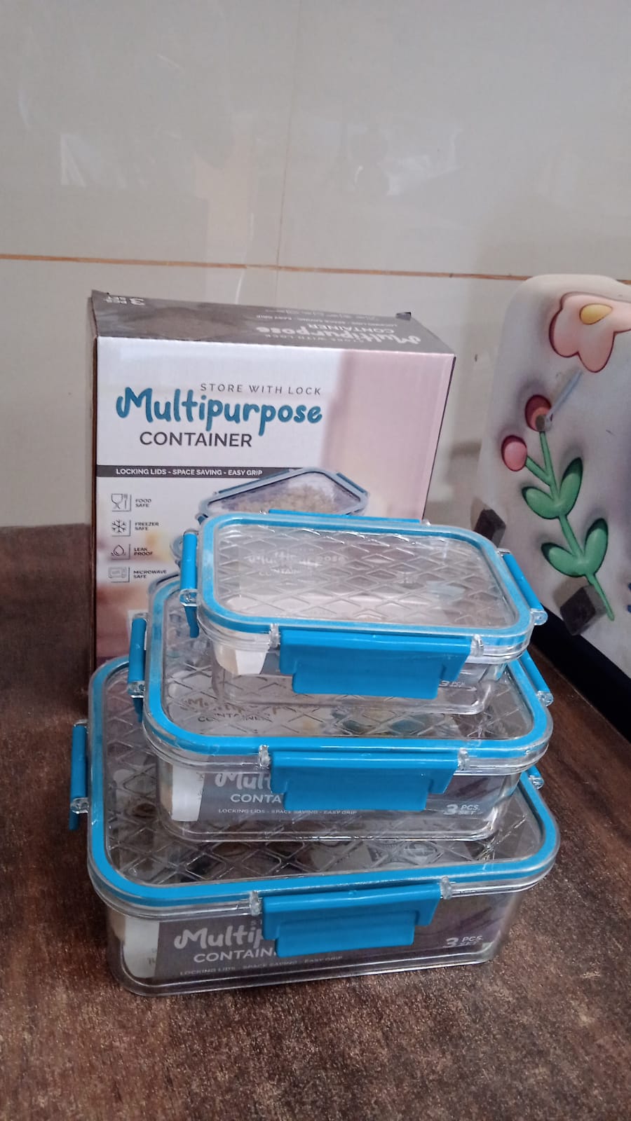 Three clear rectangular food containers with teal locking lids are stacked in front of their product box.