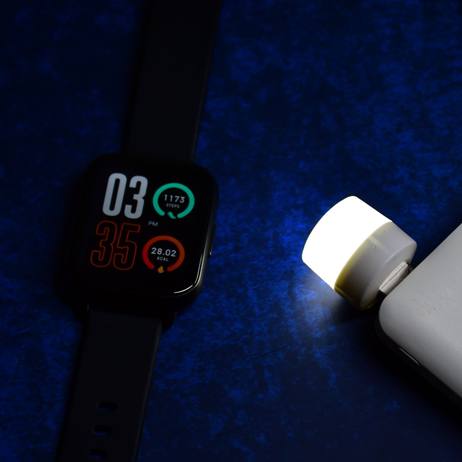 A white USB LED night light is plugged into a power bank next to a smartwatch on a dark blue surface.