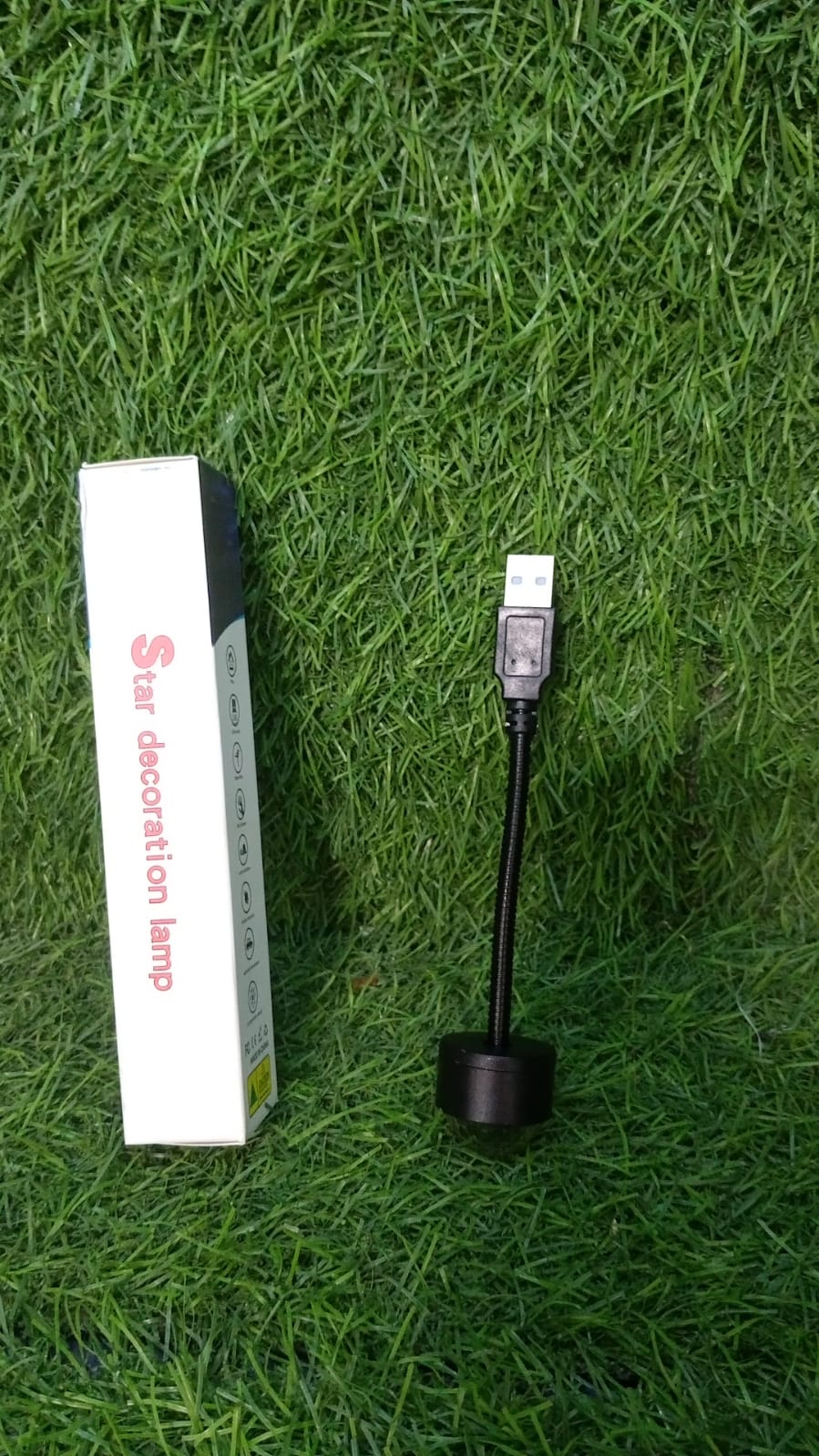 The black USB star projector and its white packaging are resting on a vibrant green grass surface.