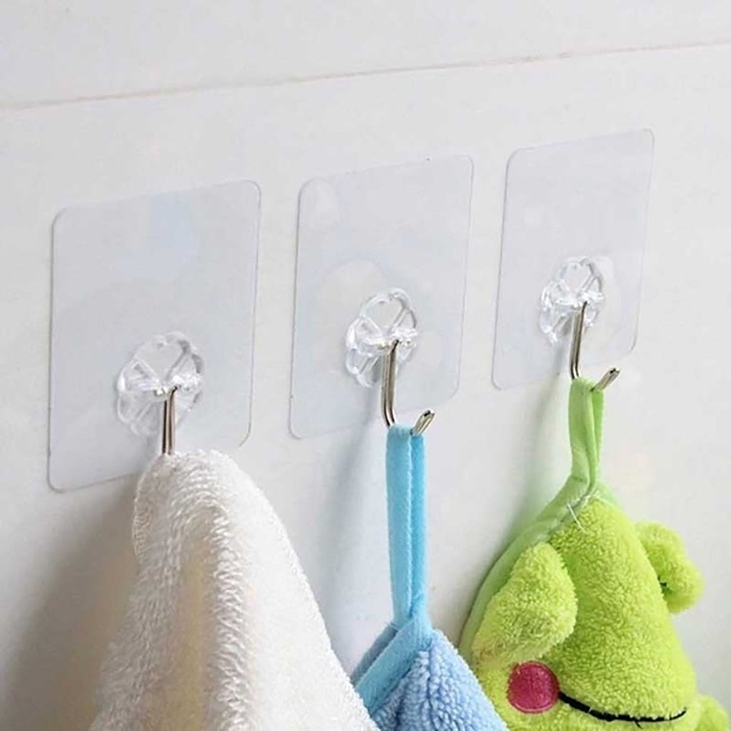 Three clear adhesive hooks with silver stainless steel hooks are mounted on a white wall, holding a white towel, a blue