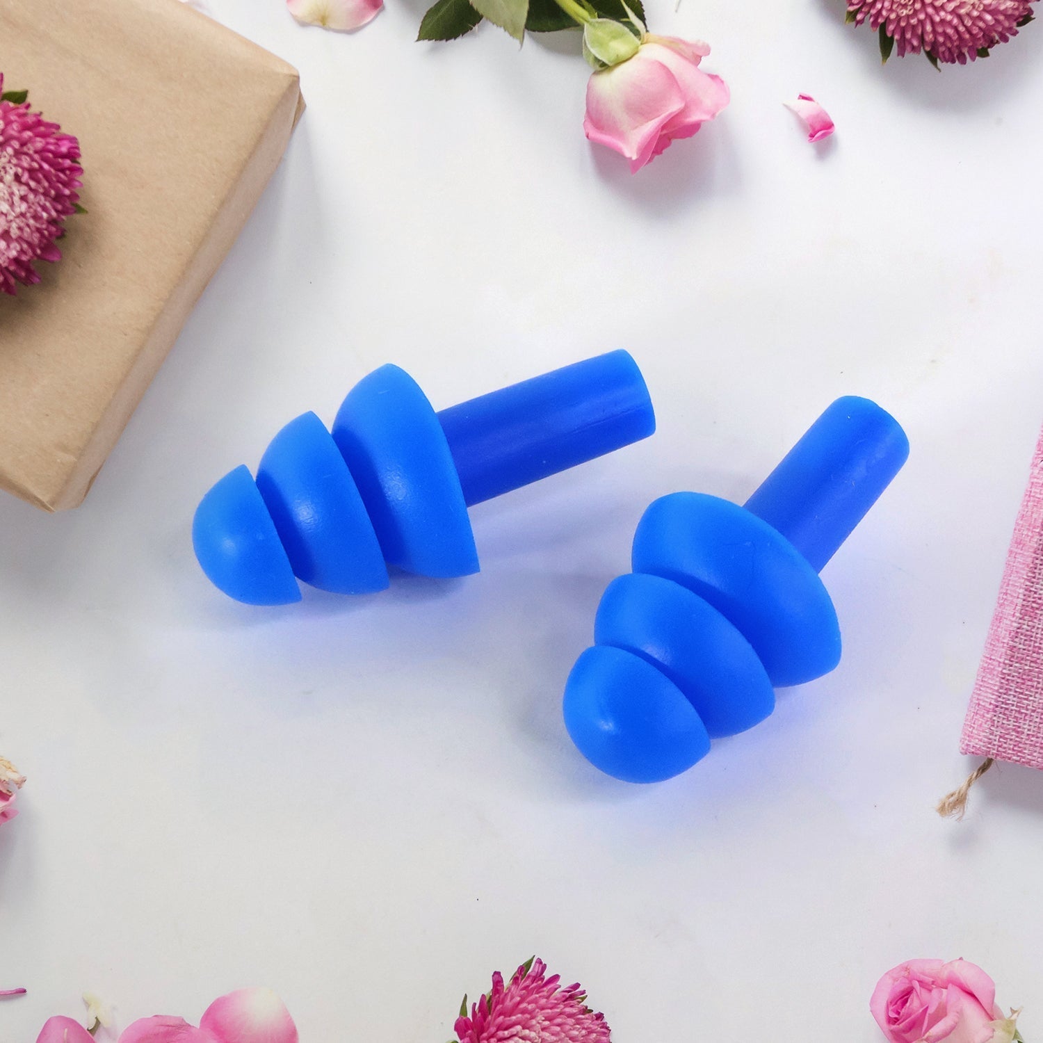 Two blue silicone earplugs are arranged on a white surface decorated with pink flowers and a brown wrapped gift.