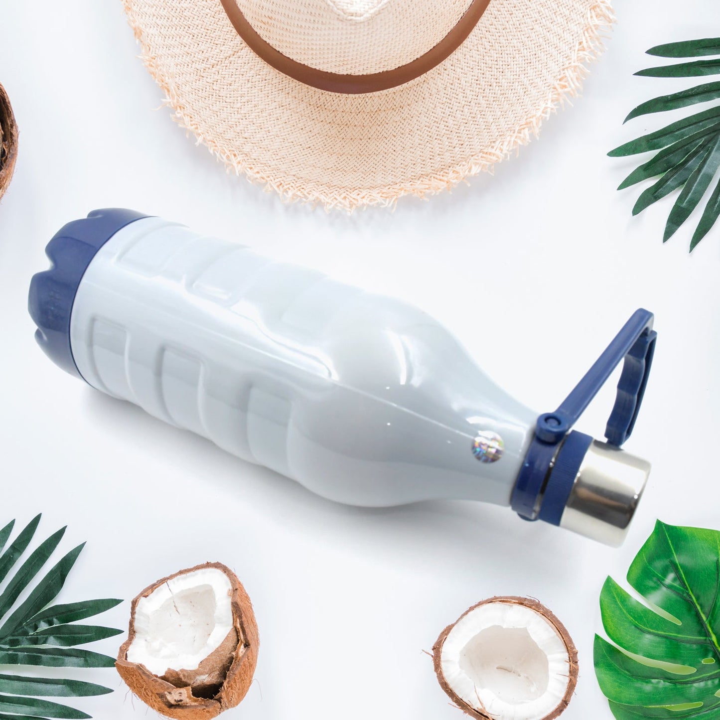 White and blue water bottle lying horizontally, surrounded by palm leaves, coconuts, and a straw hat.