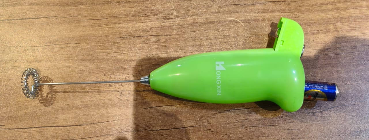 A lime green frother with its battery compartment open, revealing a blue battery, rests on a wooden surface.
