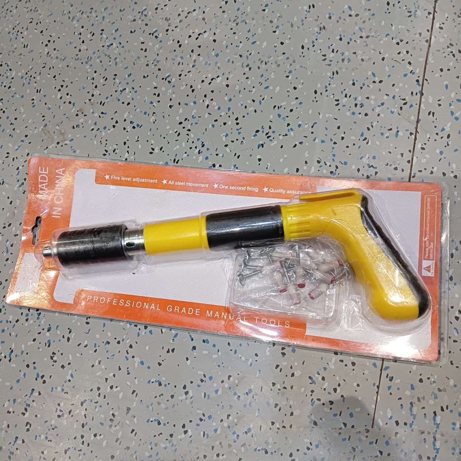 Yellow and black manual nail gun with fasteners in blister packaging, resting on a gray patterned floor.