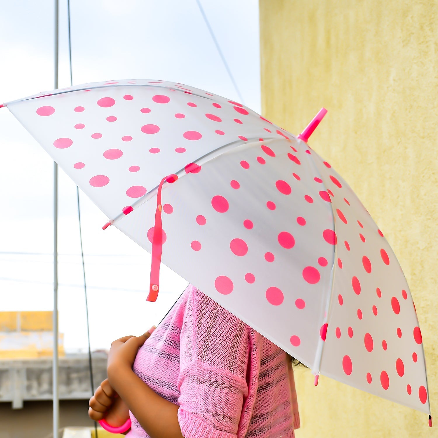 A person holds an open white umbrella with bright pink polka dots, wearing a matching pink sweater.