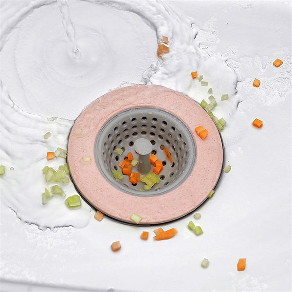 A pink and gray silicone sink strainer in a white sink, catching chopped vegetables as water flows around it.