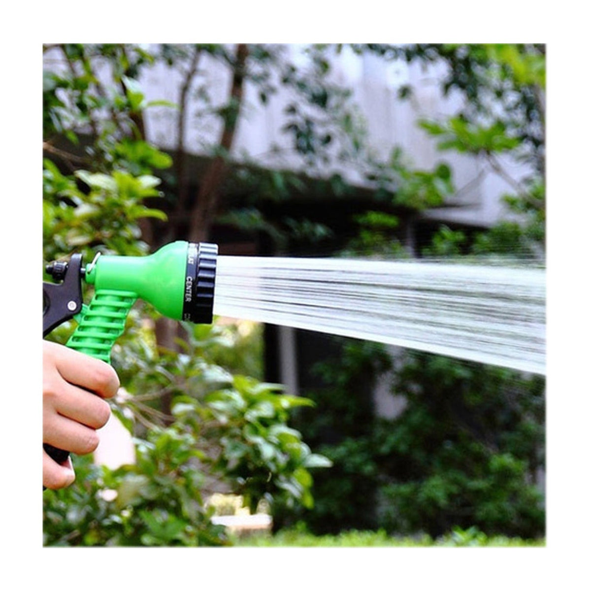 A hand holds a green and black hose nozzle spraying a strong stream of water into a blurred green garden.