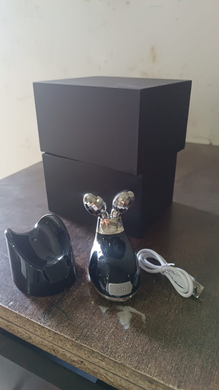 A black microcurrent massager, its charging base, a white USB cable, and two black boxes are on a wooden surface.