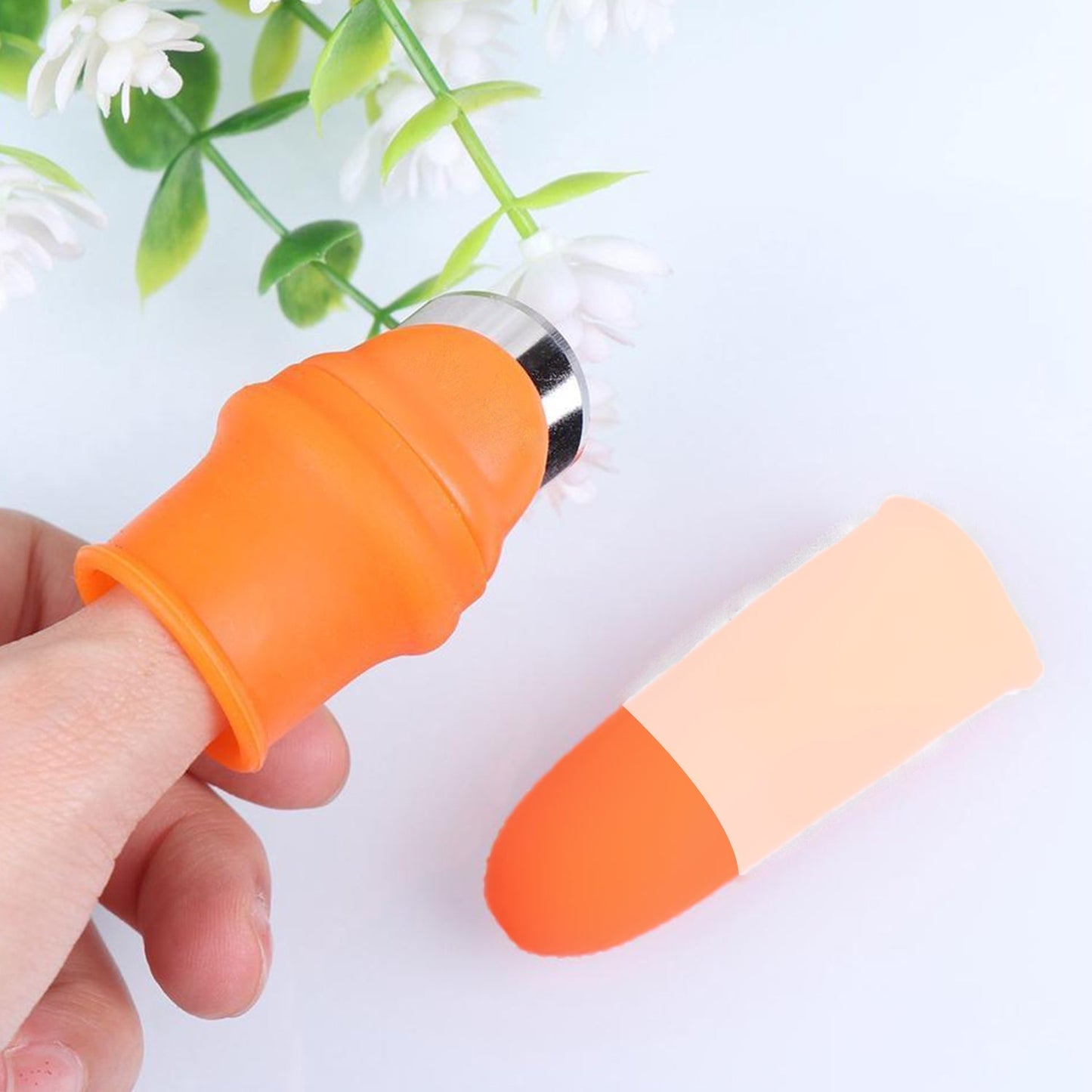 A hand wearing an orange thumb cutter with a blade, next to a beige finger protector on a white surface with a plant.