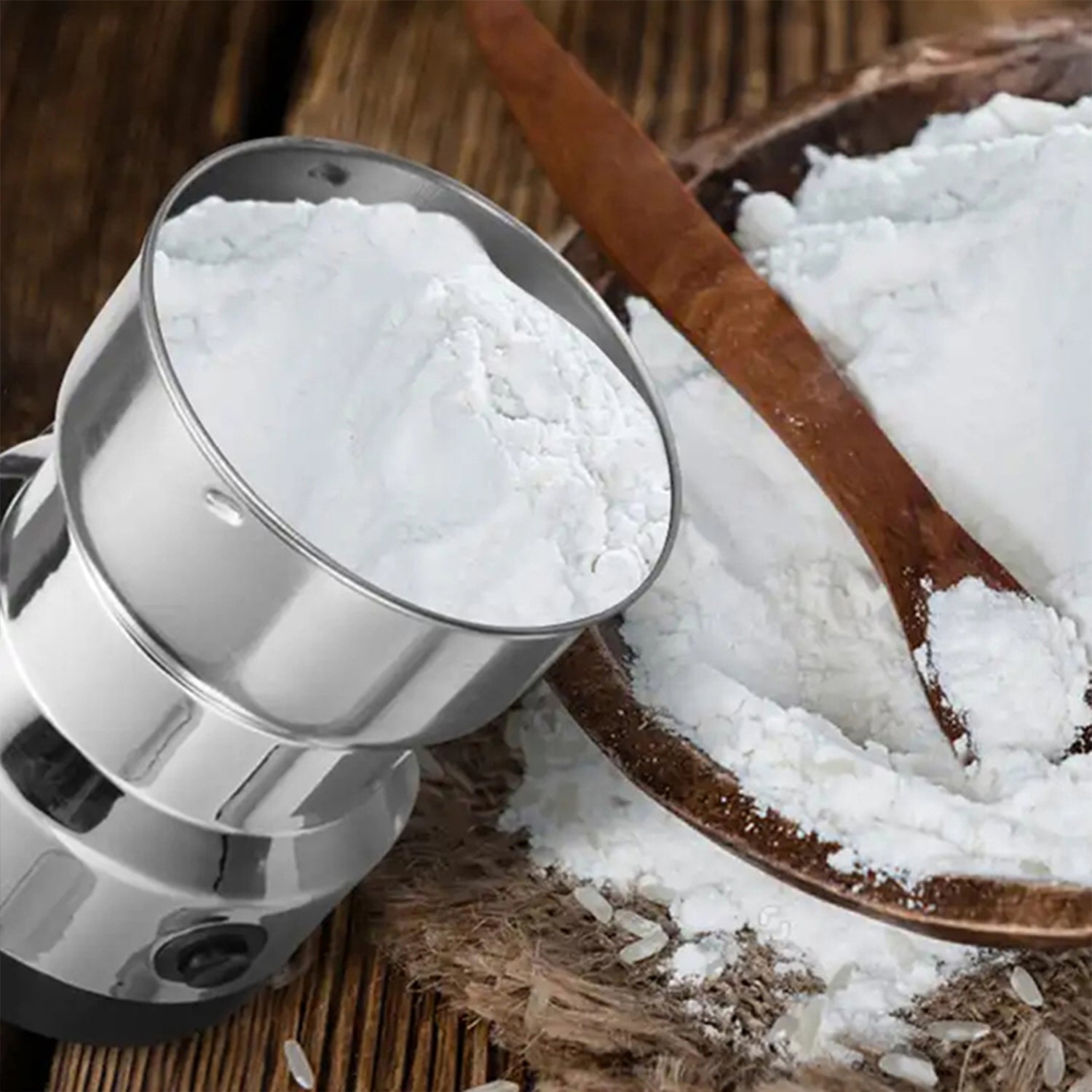 The grinder's stainless steel bowl is filled with white flour, next to a wooden bowl of flour and a spoon.