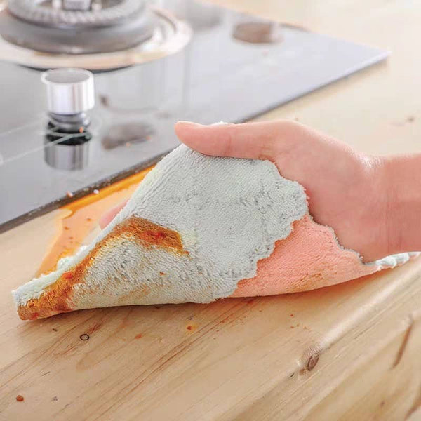 A hand holds a light blue and pink dual-sided towel, wiping a spill from a wooden kitchen countertop.