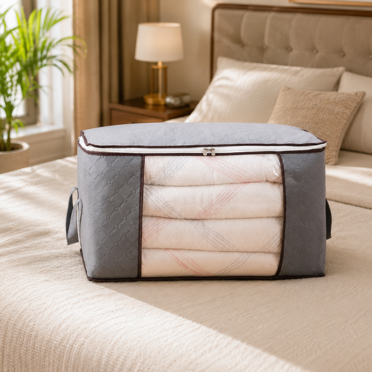 A grey quilted fabric storage bag with a clear window filled with folded white blankets sits on a bed.