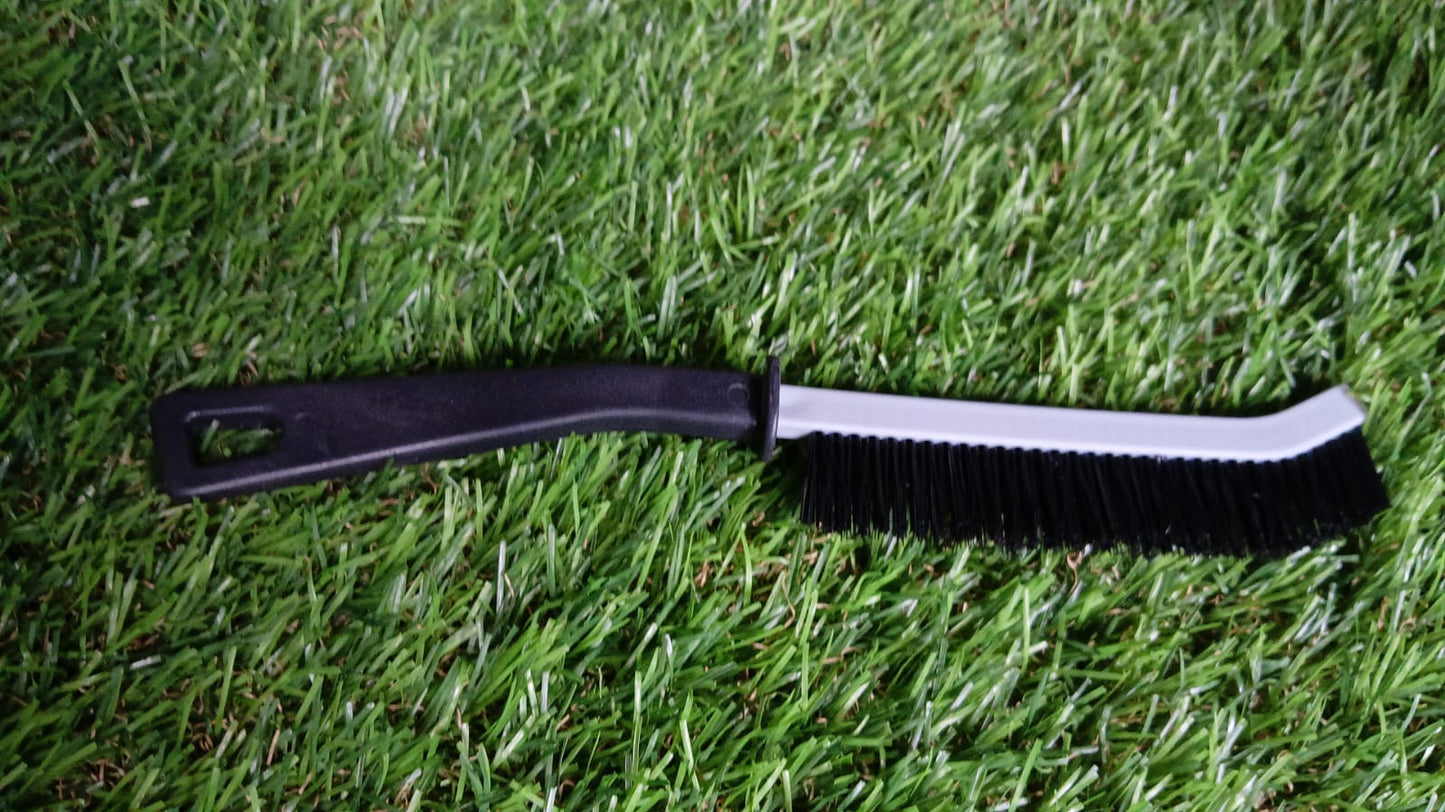 A black and gray crevice cleaning brush with black bristles rests horizontally on a vibrant green artificial grass surface.