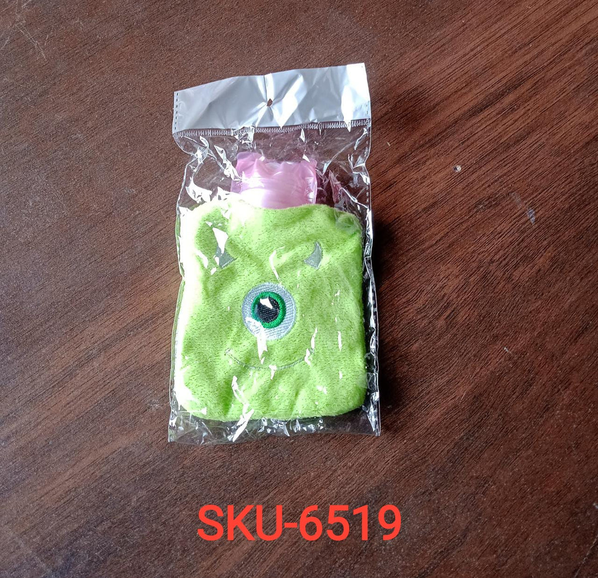 A green monster hot water bag with a pink top is sealed in clear plastic packaging on a wooden surface.