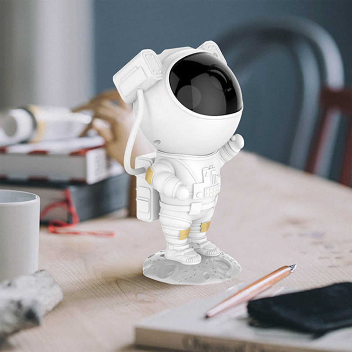 A white astronaut projector stands on a wooden desk, surrounded by books and a pen, with a white mug to the left.
