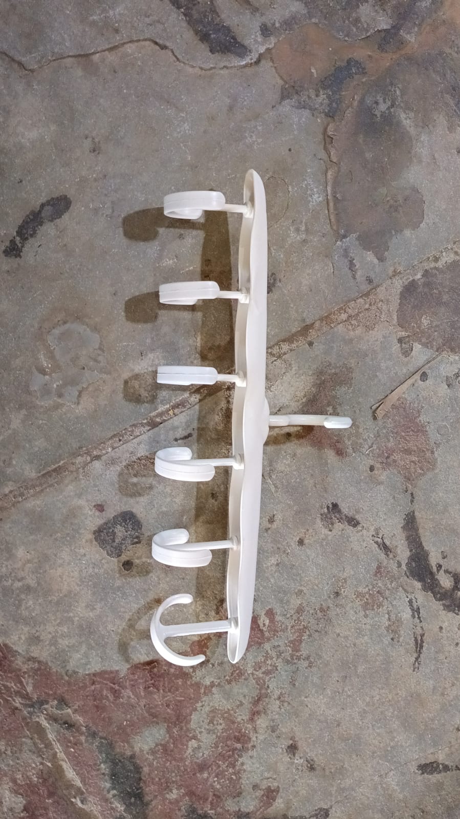 A white plastic multi-hook hanger with six rotating hooks, lying flat on a textured grey and brown surface.