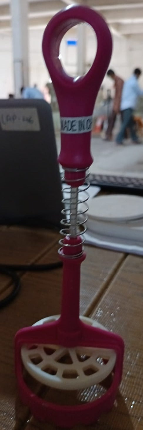A pink and white potato masher stands on a wooden table, with people and a laptop blurred in the background.