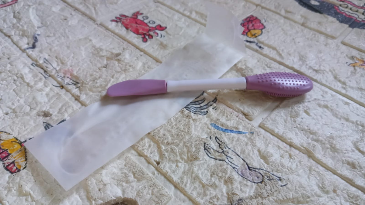 A purple and white double-headed silicone mask brush resting on its clear plastic packaging on a patterned surface.