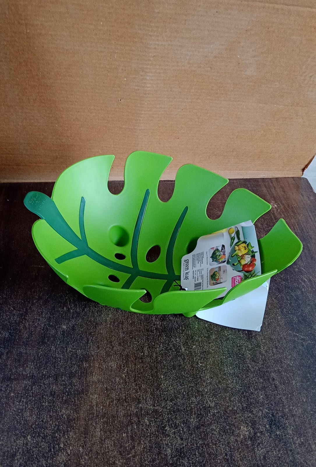 A green leaf-shaped plastic fruit basket with a product label inside, sitting on a dark wooden surface.