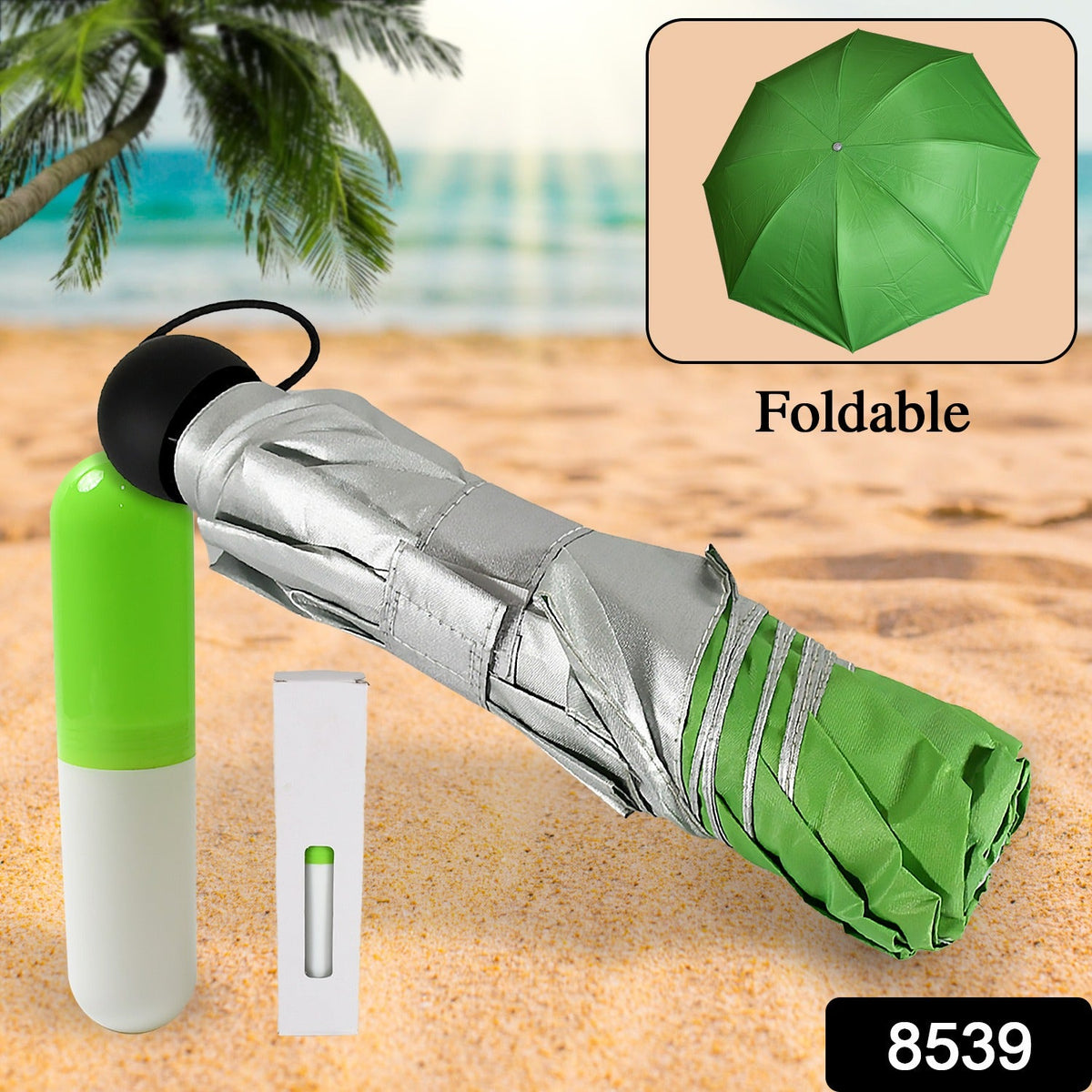 A folded green and silver umbrella, its capsule case, and a box lie on a sandy beach with palm trees and ocean.