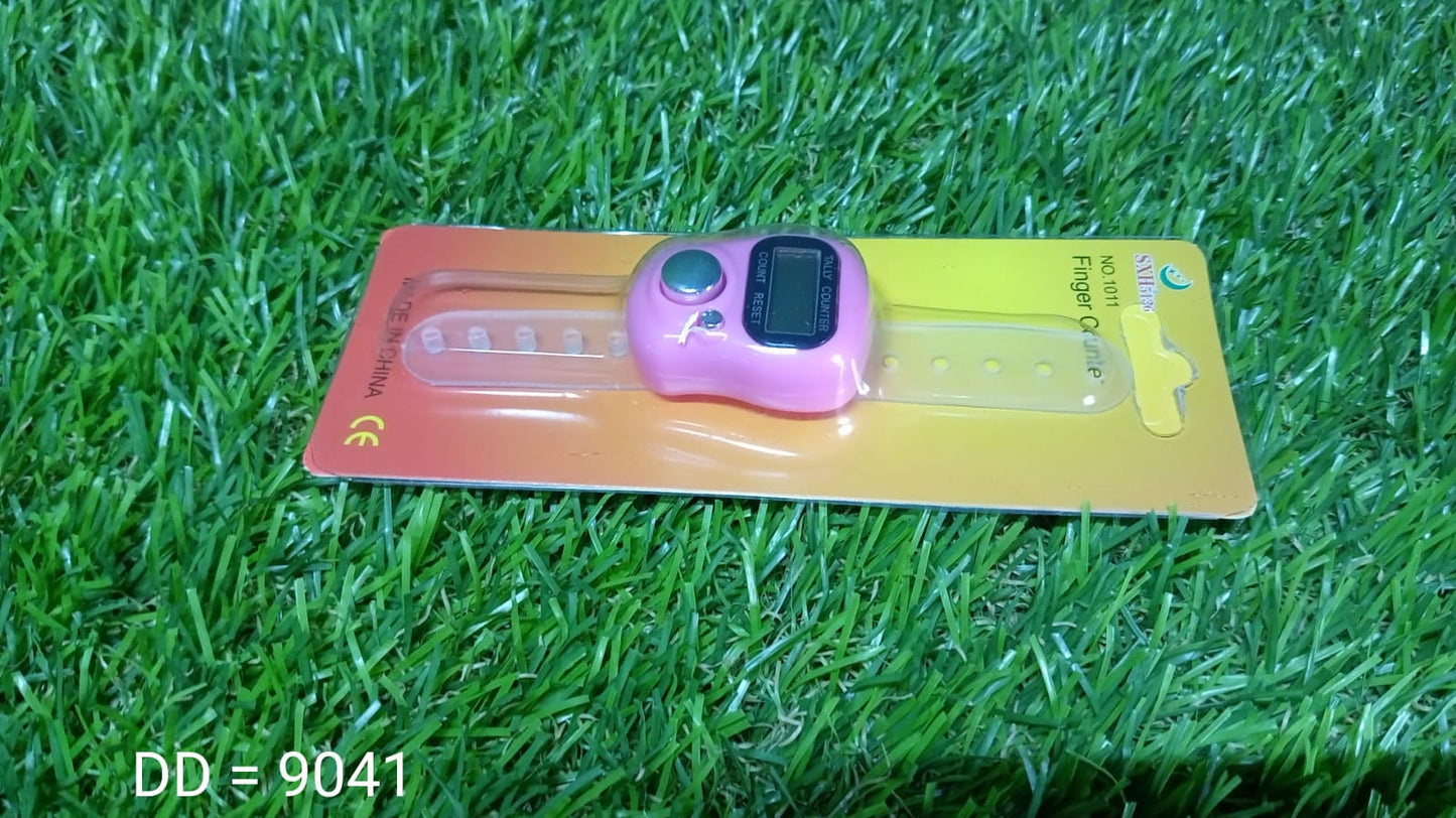 A pink digital tally counter with a clear strap is packaged on a yellow and orange card, lying on green grass.