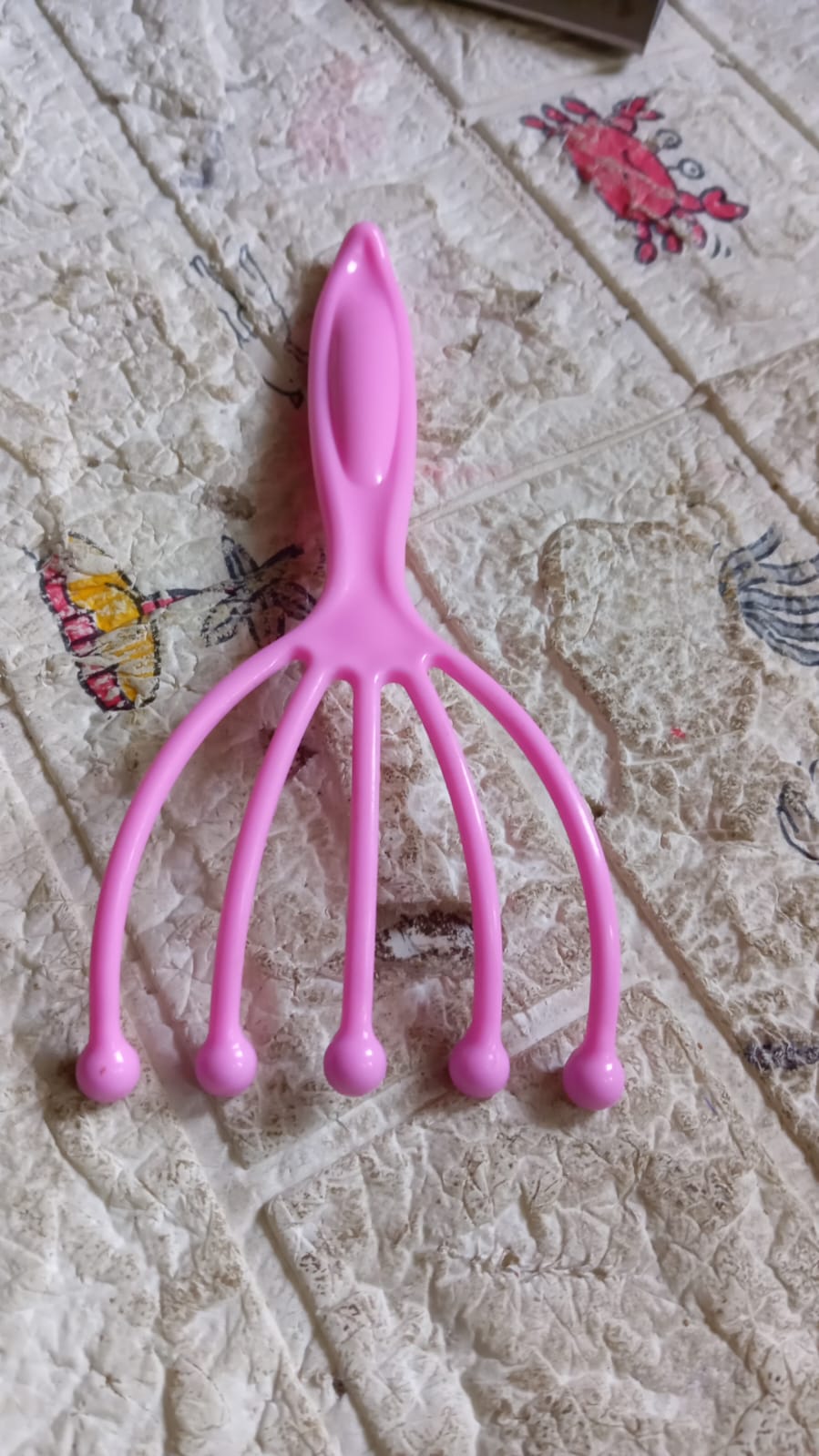 A pink plastic scalp massager with five flexible arms ending in rounded pink tips, lying on a textured white and beige