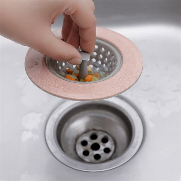 A hand lifting a pink and gray silicone sink strainer filled with chopped vegetables from a stainless steel sink drain.