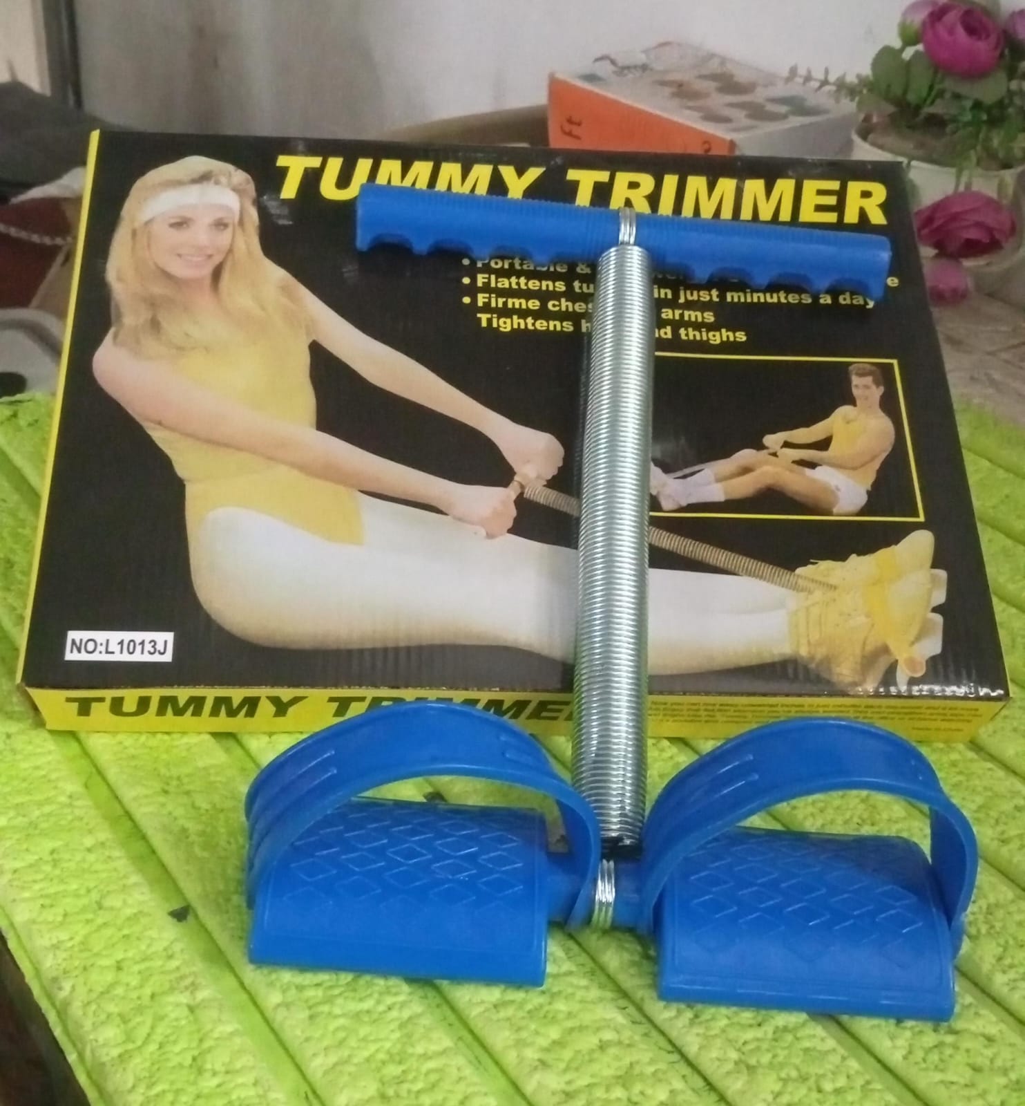 A blue foot pedal exerciser with a metal spring sits in front of its yellow and black product box on a green textured