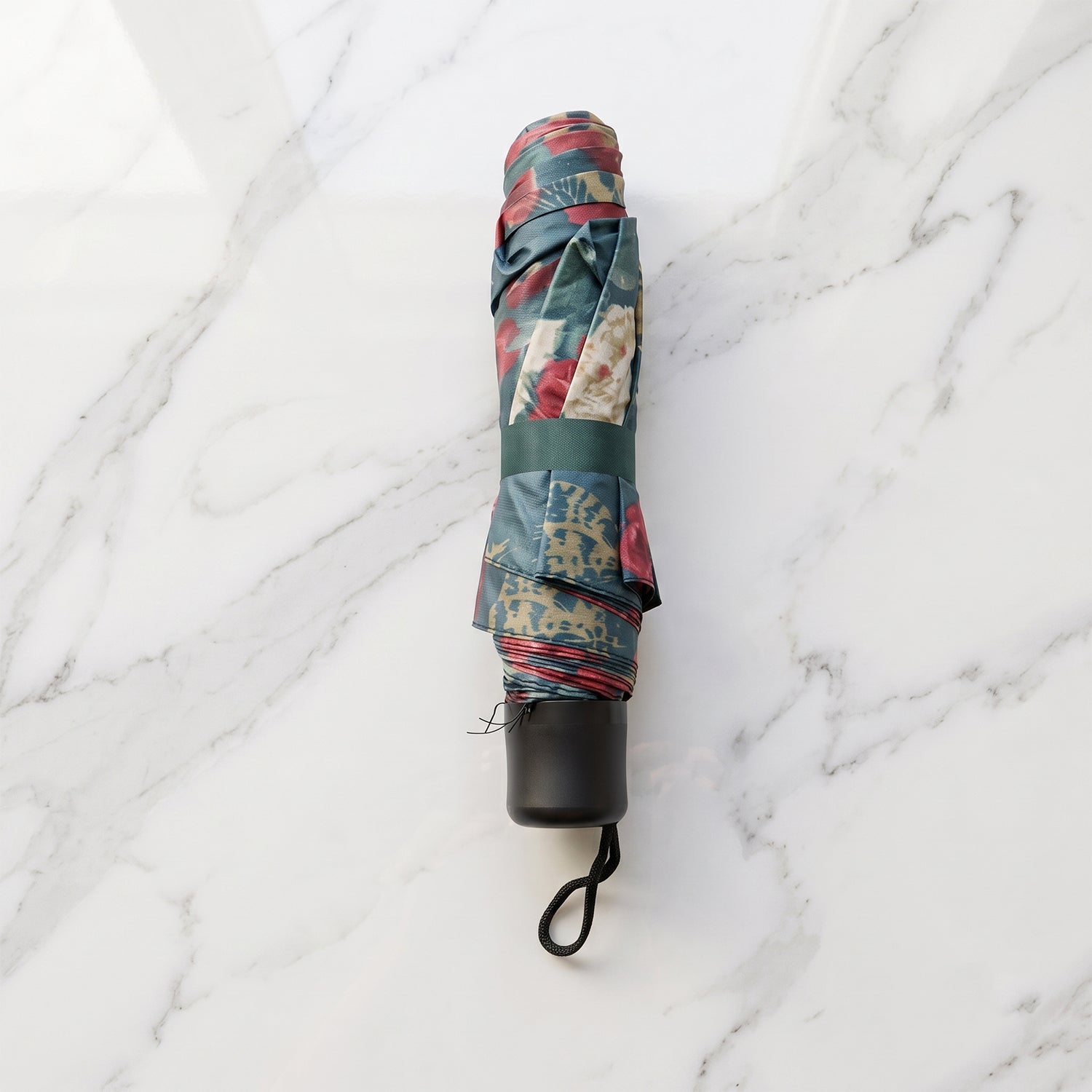 A folded floral umbrella, secured with a dark green strap, rests on a white marble surface, showing its black handle and