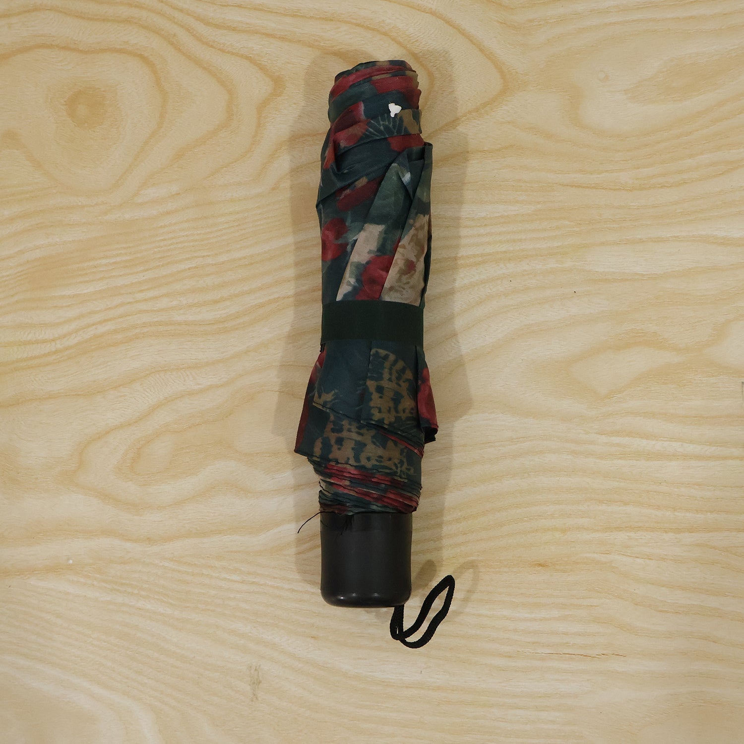 A folded floral umbrella, secured with a dark green strap, rests on a light wood surface, showing its black handle and wrist