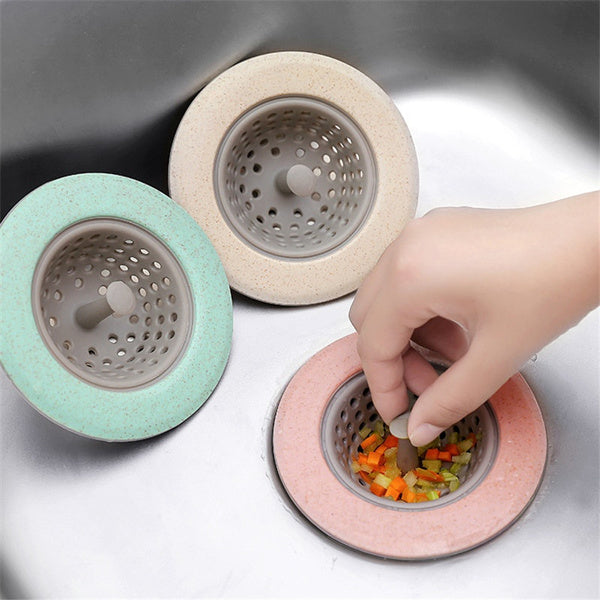 Three sink strainers in mint green, beige, and pink are in a stainless steel sink, with a hand placing food in the pink one.