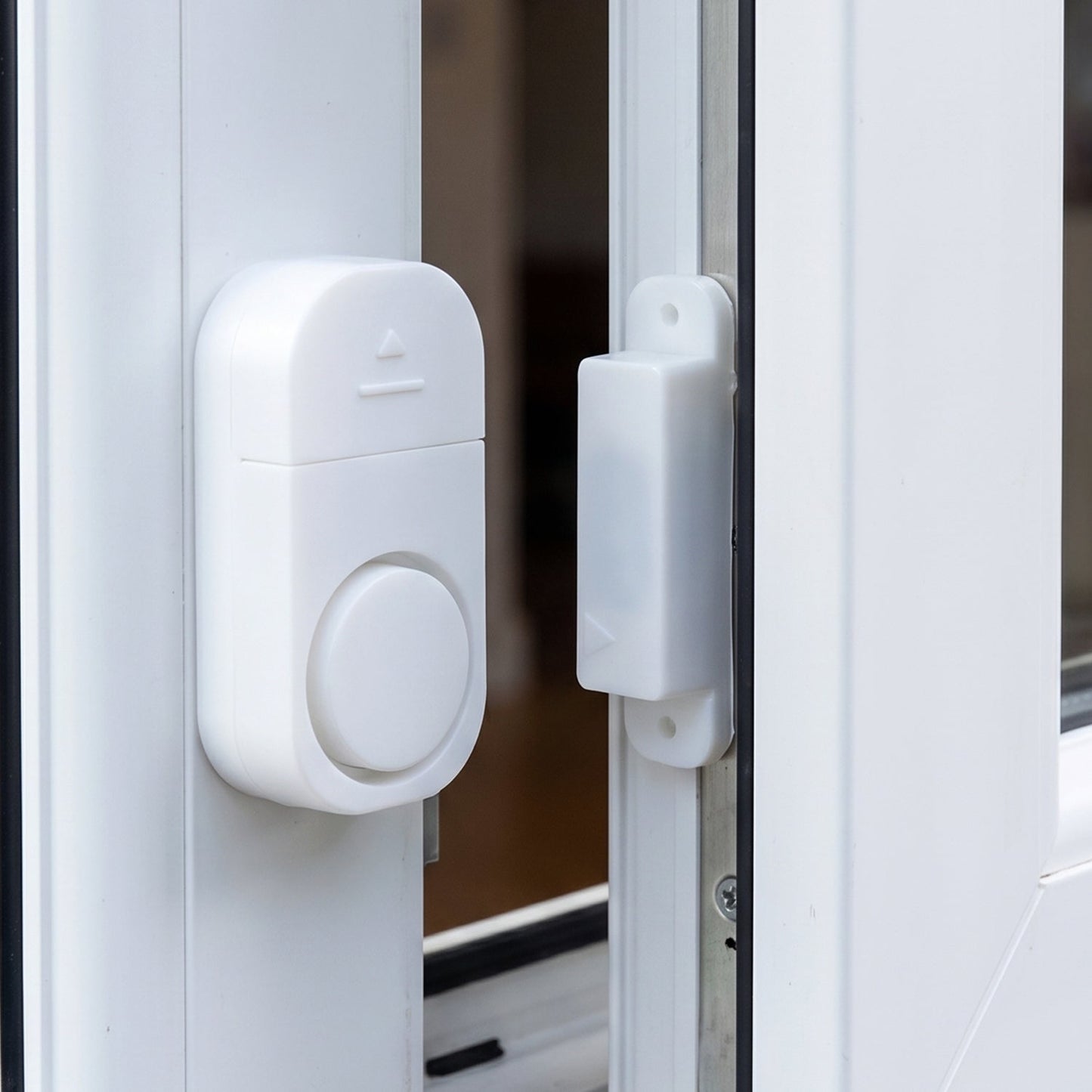 A white plastic magnetic alarm is attached to a white window frame, with its smaller magnetic component on the adjacent