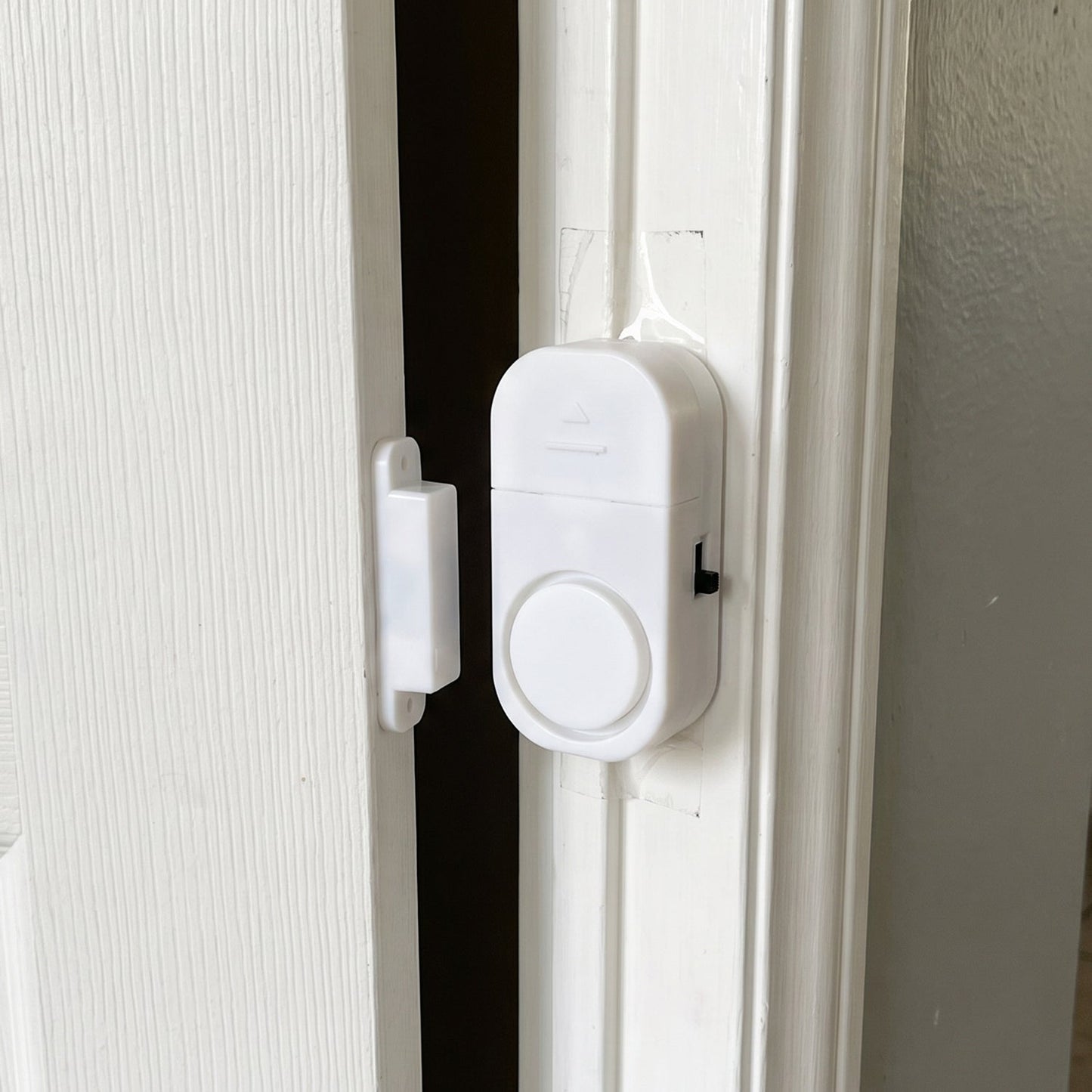 A white magnetic door alarm is installed on a slightly ajar white door frame, with the two parts separated.