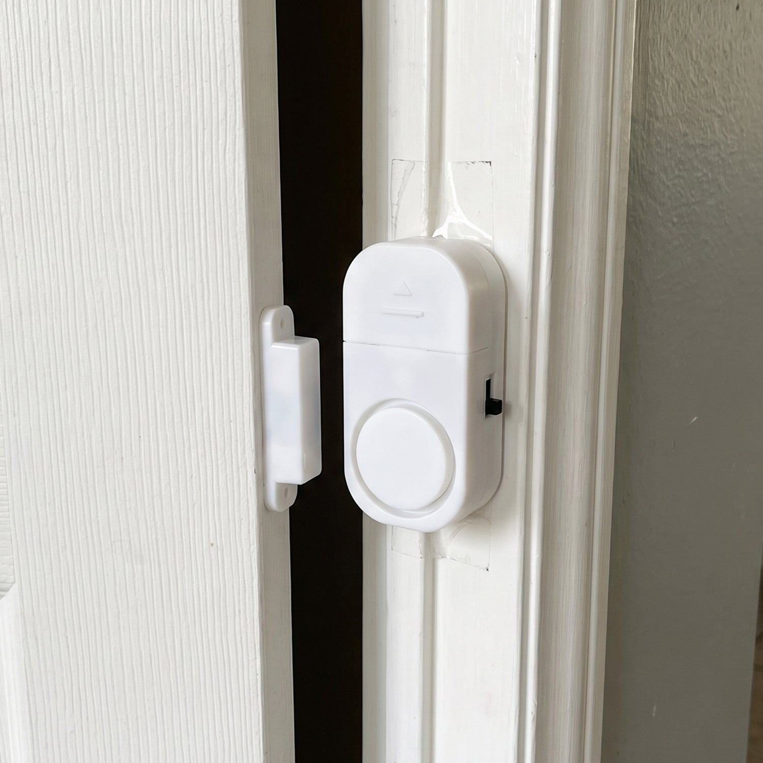 A white magnetic door alarm is installed on a slightly ajar white door frame, with the two parts separated.