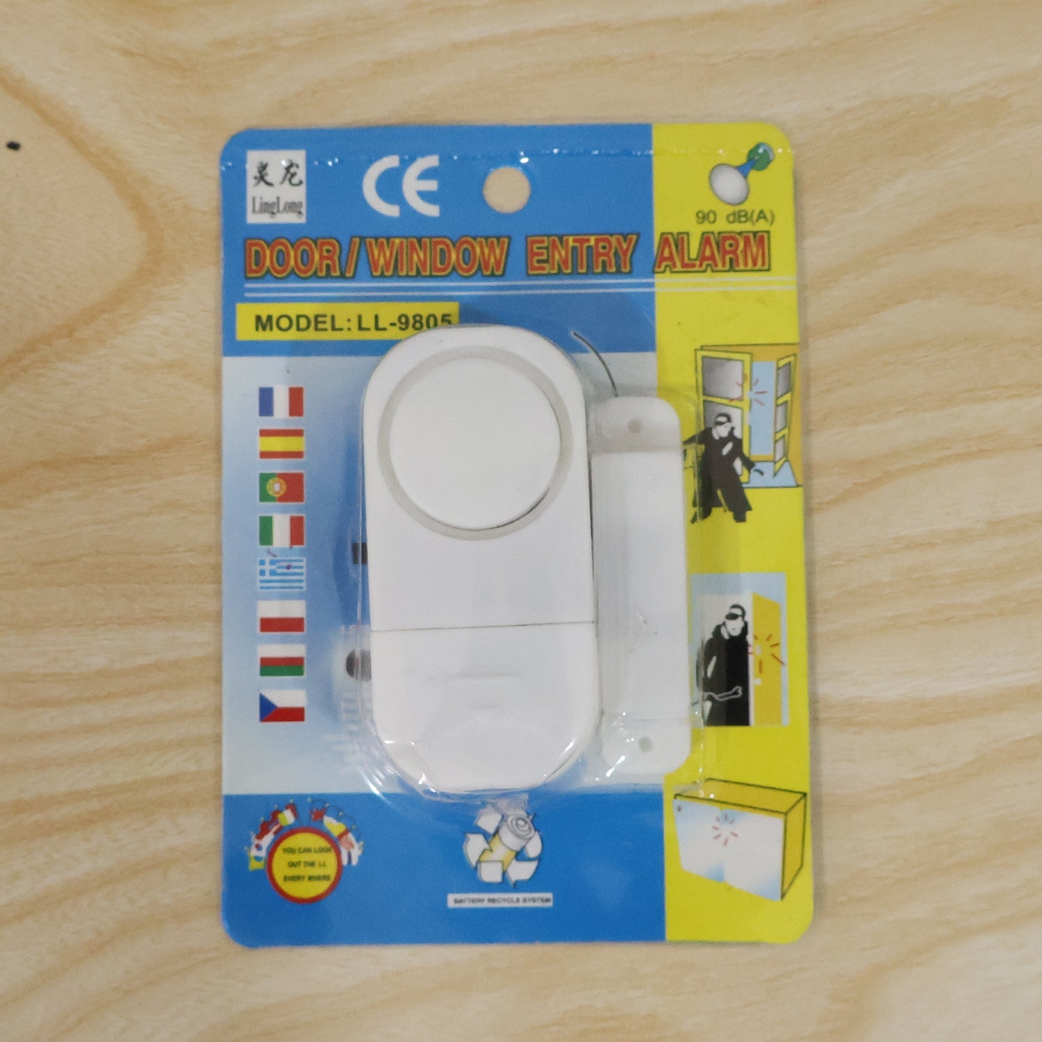 A white magnetic door/window entry alarm is sealed in its blue and yellow retail packaging on a light wooden surface.